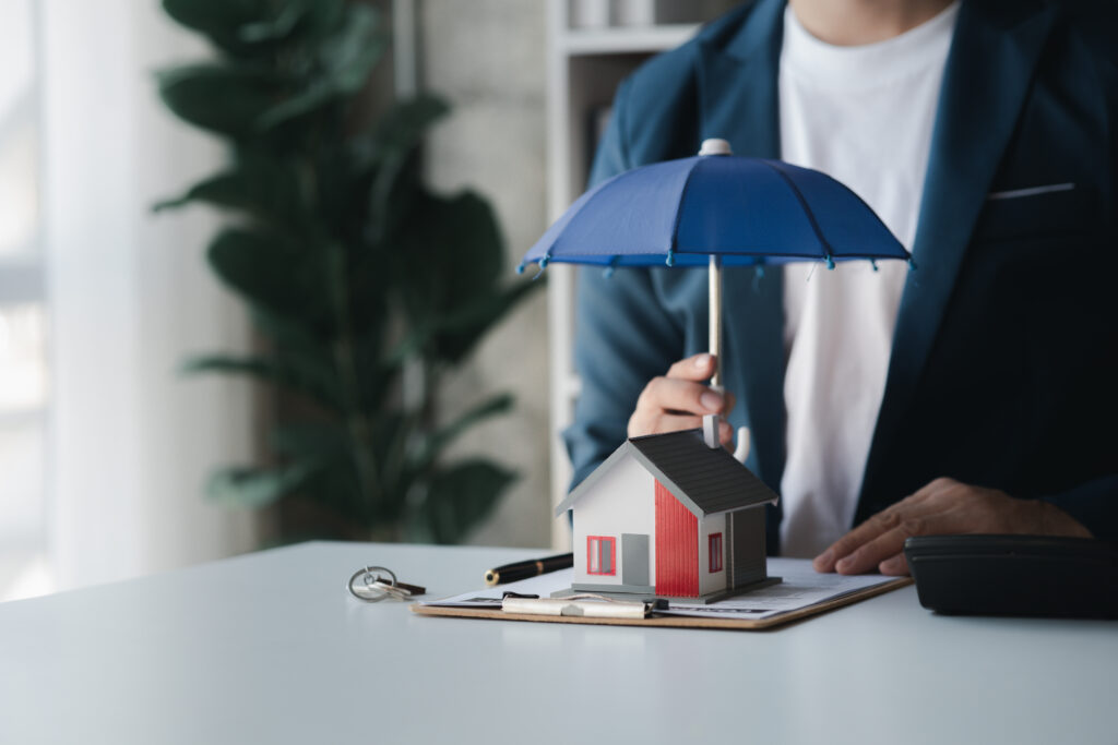 Person holding umbrella over miniature house symbolizing home protection.
