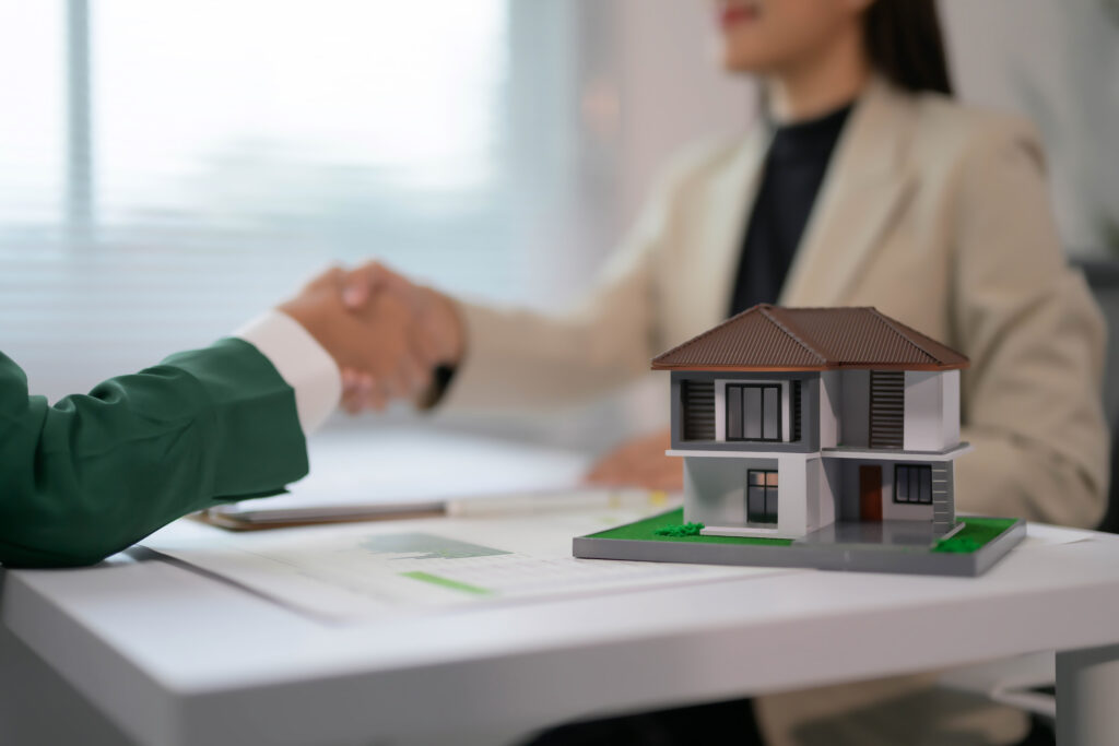 Two people shaking hands over a house model and documents.