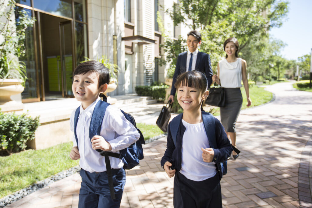 Two children walking to school with backpacks, followed by two adults.