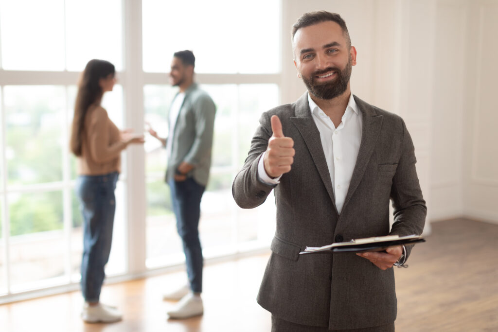 Confident real estate agent giving thumbs up with couple discussing in background.