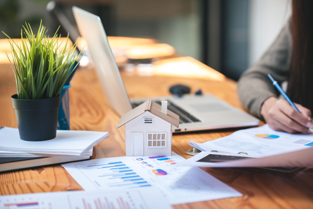 A small model house on financial documents with a laptop in the background.